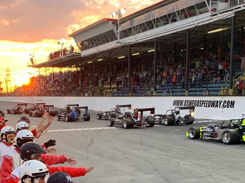 Photo of Novelis Supermodifieds at Oswego Speedway courtesy of Oswego Speedway. Packed granstands overlook the front strecth, and pit crew workers are lined up